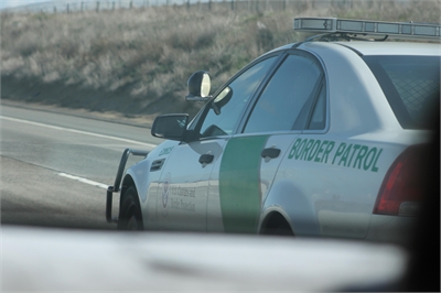 [ai] A U.S. Border Patrol vehicle parked on the side of a highway. The car features a green stripe and the words 'BORDER PATROL' on the side, with a blurred view of another vehicle in the foreground.